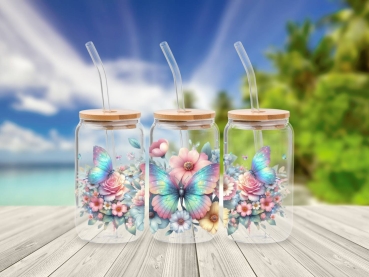 Drei 16 oz Glasbecher mit Bambusdeckel und Glastrinkhalm, verziert mit lebendigen Blumenmotiven und schillernden Schmetterlingen in Pastelltönen. Die Becher stehen auf einem Holztisch vor einem tropischen Strandhintergrund, was eine fröhliche und naturver